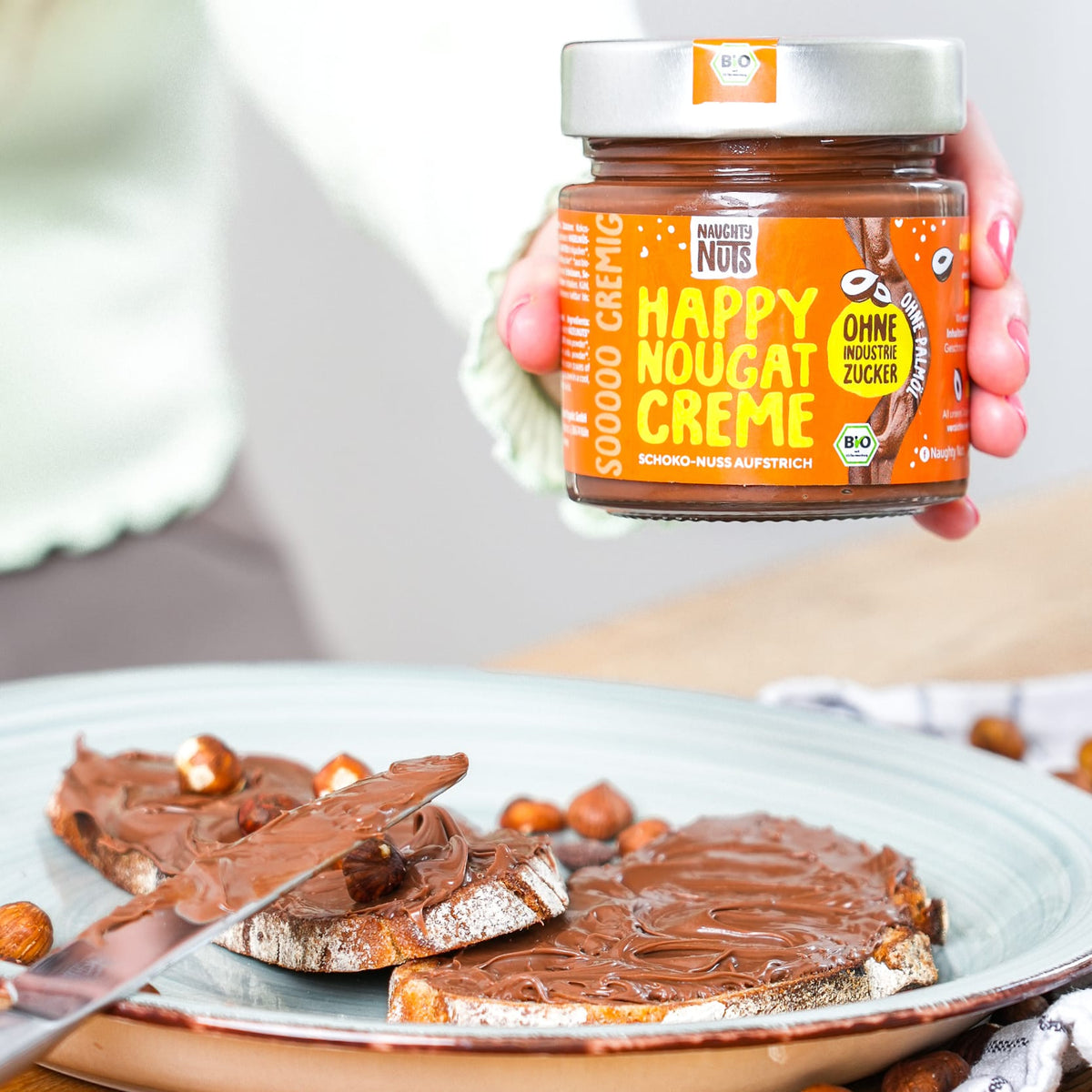 Hand hält Naughty Nuts Happy Nougat Creme Glas über zwei Brotscheiben mit Aufstrich und Haselnüssen