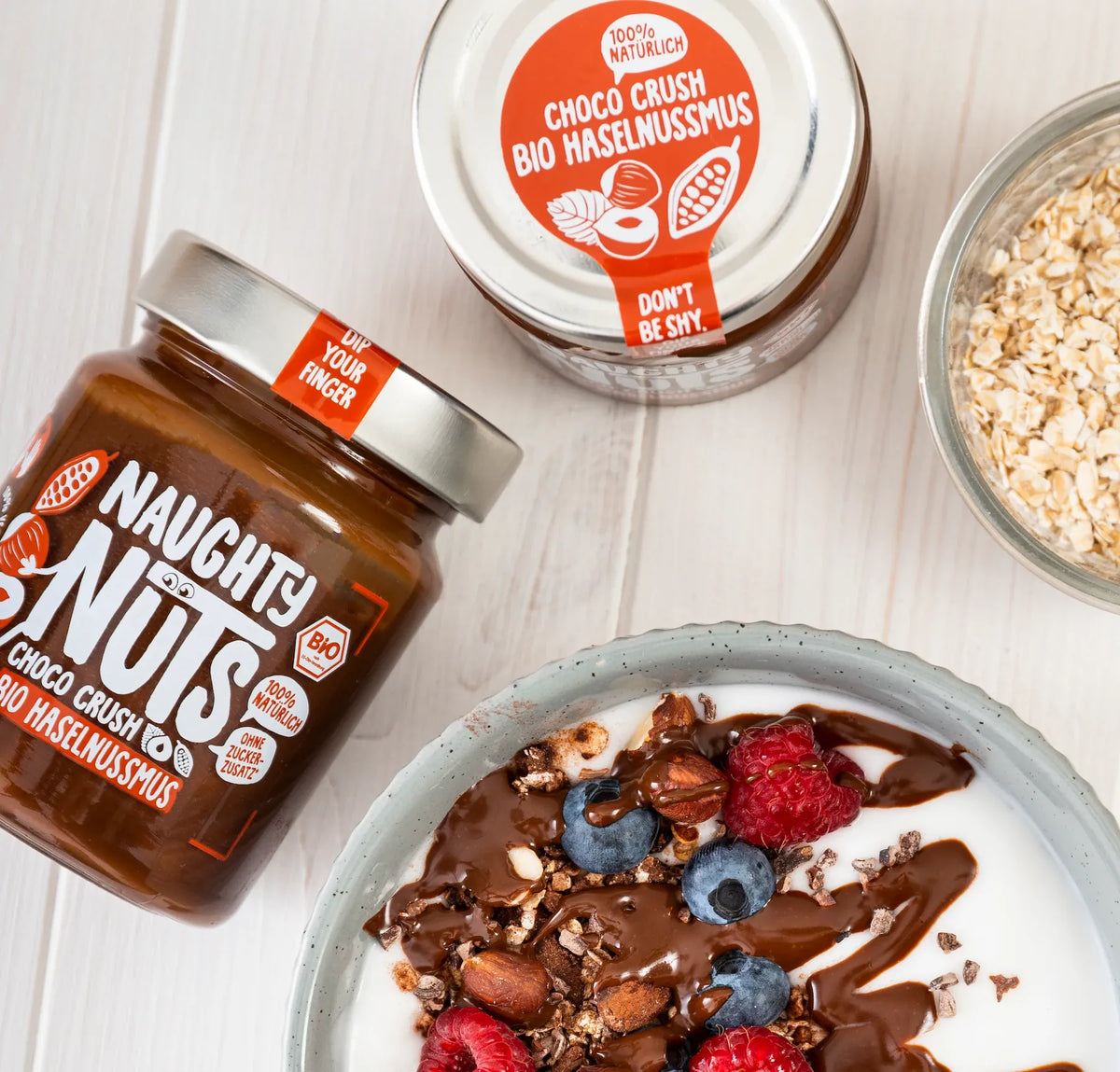 Zwei Gläser Naughty Nuts Choco Crush Bio Haselnussmus, eine Schale Müsli mit Beeren und Haferflocken