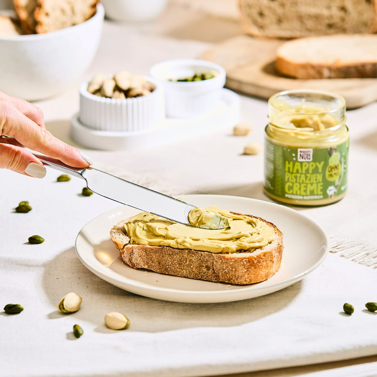 Hand streicht Happy Pistazien Creme auf eine Scheibe Brot, daneben ein Glas Pistaziencreme und Pistazien
